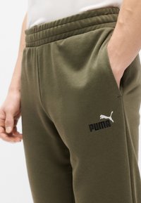 Φόρμες σε ελιά πράσινο χρώμα από απαλό ύφασμα, με ελαστική μέση, πλαϊνές τσέπες και μαύρο λογότυπο PUMA με λευκή σιλουέτα.