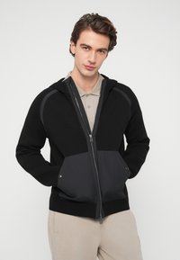 Μαύρο μπουφάν με κουκούλα και φερμουάρ, κατασκευασμένο από υφασμάτινη πλέξη με υφή και στερεή μαύρη λεπτομέρεια στη μέση. Διαθέτει γκρι λεπτομέρειες και μπροστινές τσέπες.