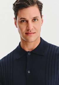 Młody mężczyzna z krótkimi brązowymi włosami, jasnymi oczami i lekko uśmiechnięty nosi ciemnoniebieską koszulę z guzikami na tle jednolitego białego tła.