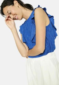 Zilā ziedu raksta blūze ar viļņainām piedurknēm, teksturēts audums, kombinēta ar baltām nūtām biksēm. Tuvplāns uz modeļa augšdaļu.