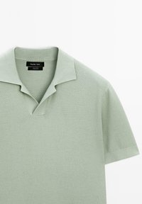 Polo shirt in cotone verde chiaro con design testurizzato, presenta un colletto classico, maniche corte e un'apertura a V. Etichetta visibile sul colletto.