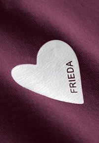 Bordeaux stof met een gewoven witte hartdetail waarop het woord "FRIEDA" in diep maroon is gedrukt. Een gladde oppervlakte en contrasterende kleuren.