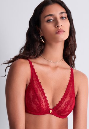 Jeune femme aux longs cheveux foncés portant un soutien-gorge en dentelle rouge, un collier en or et des boucles d'oreilles en or, sur un fond uni.