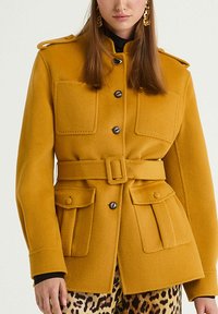 Veste en laine jaune moutarde avec un design inspiré de l'armée, grandes poches avant, taille ceinturée et fermetures à boutons.