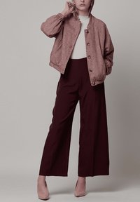 Chaqueta bomber rosa con patrón de pata de gallo y cierre de botones, combinada con pantalones oscuros de talle alto. Modelo lleva una blusa clara y tacones beige.