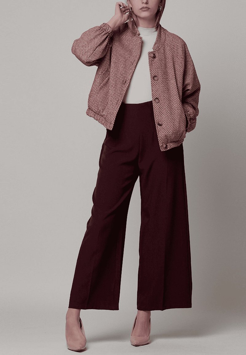 Chaqueta bomber rosa con patrón de pata de gallo y cierre de botones, combinada con pantalones oscuros de talle alto. Modelo lleva una blusa clara y tacones beige.