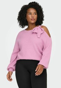Rosa gestrickter Pullover mit Cold-Shoulder-Design, geripptem Muster und einer großen Schleifenapplikation auf der linken Schulter. Dazu werden schwarze Hosen getragen.