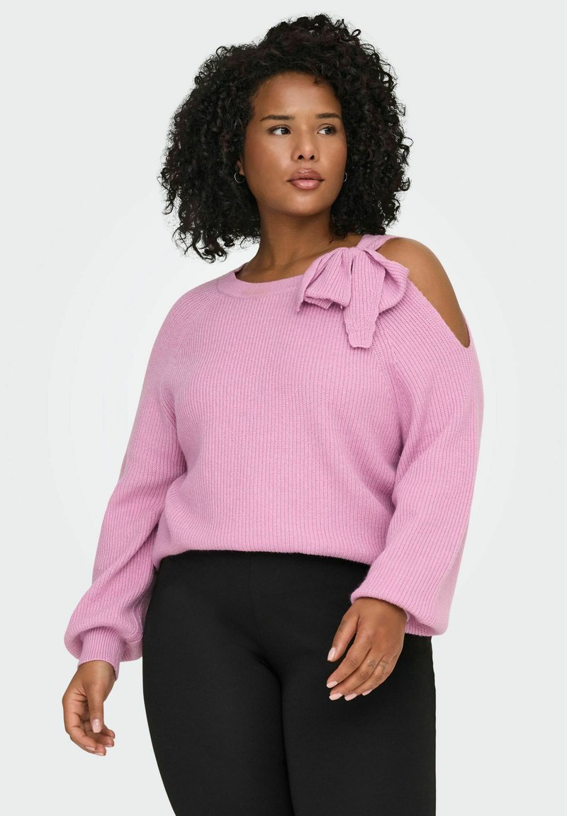 Rosa gestrickter Pullover mit Cold-Shoulder-Design, geripptem Muster und einer großen Schleifenapplikation auf der linken Schulter. Dazu werden schwarze Hosen getragen.