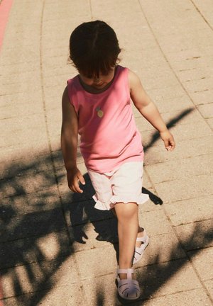 Tout-petit portant un haut sans manches rose et un short, marchant sur un chemin pavé baigné de lumière avec des ombres projetées autour.