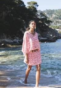 Roza haljina za plažu s uzorkom, labavim rukavima, strukirana u struku i s kontrastnim remenom; model nosi nakit i stoji pokraj vode.