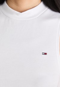Top sin mangas blanco con un cuello alto y un pequeño logo bordado en rojo, blanco y azul en el lado izquierdo del pecho.