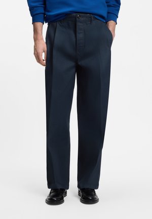 Mann trägt dunkelblaue, gerade geschnittene Hose mit Seitentaschen und schwarze Anzugschuhe, steht mit teilweise in den Taschen befindlichen Händen.