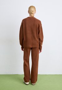 Braunes Strickpullover- und Hosen-Set mit lockerer Passform, weichem Stoff und gerippten Bündchen, vor einem hellen Hintergrund und einem grünen Boden.