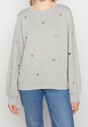 Femme portant un sweat-shirt gris clair à manches longues avec une petite broderie fleurie colorée, associée à un jean bleu, debout devant un fond uni.