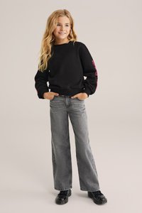 Melna džemperis ar rozā grafiskajiem akcentiem uz piedurknēm, apvienots ar pelēkiem platajiem džinsiem un melnām kurpēm. Ikdienišķs, brīvs piegriezums un mīksta tekstūra.