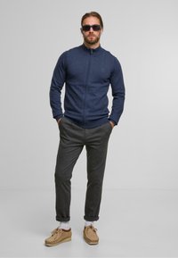 Marineblaue Zip-Up-Strickjacke in strukturiertem Knit, kombiniert mit grauen schmal zulaufenden Hosen und hellbraunen Schnürslippers. Als Accessoires dienen dunkle Sonnenbrillen.