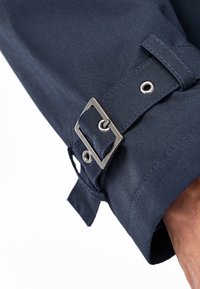 Manga azul marino con un hebilla rectangular de metal, una correa de tela y un ojal plateado, que enfatiza un diseño estructurado y a medida.