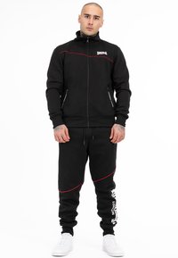 Zwarte trainingsjack met rits en rode accenten, gecombineerd met zwarte sweatpants met rode piping en witte logodetail, gedragen met witte sneakers.