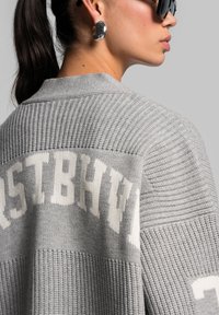 Gray knitted cardigan with textured stripes, featuring white lettering on the back. Solid color with a relaxed fit and wide sleeves.