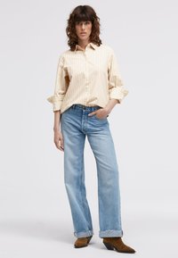 Beige button-up shirt with vertical stripes, paired with light blue straight-leg jeans and brown heeled boots, standing against a light background.