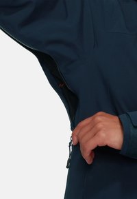 Navy waterproof jacket with a zippered vent under the left arm. Smooth fabric texture and adjustable cuff detail visible.
