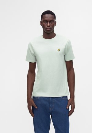 Homme portant un t-shirt vert clair à manches courtes avec un petit logo oiseau jaune sur la poitrine et un jean bleu en denim, debout sur fond blanc.