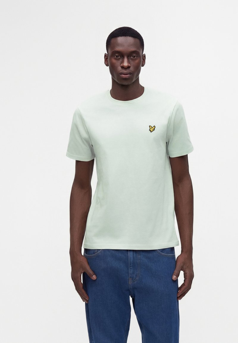 Homme portant un t-shirt vert clair à manches courtes avec un petit logo oiseau jaune sur la poitrine et un jean bleu en denim, debout sur fond blanc.