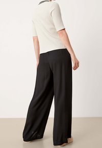 Hladký krémový top s krátkým rukávem a černými lemy, spárovaný s volnými černými kalhotami, model z pohledu zezadu, neutrální pozadí.