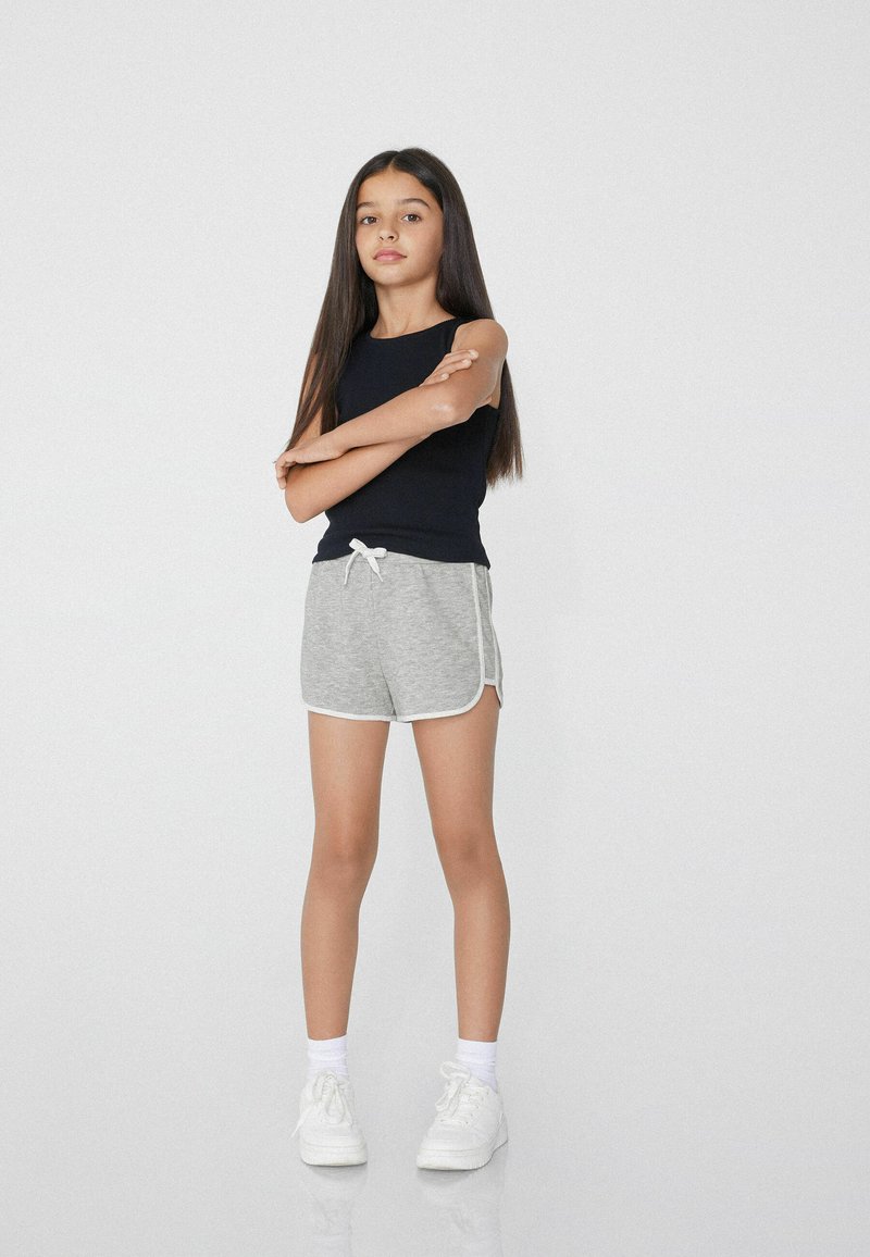 Canottiera nera, pantaloni corti grigi con rifiniture bianche e sneakers bianche. I pantaloni corti sono dotati di cintura e hanno una vestibilità comoda, mostrati su uno sfondo neutro.
