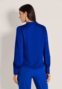 Blaue Bluse mit langen Ärmeln, aus glattem Stoff, mit Kragen und geknöpften Manschetten. Hat einen abgerundeten Saum und ist mit passenden blauen Hosen kombiniert.