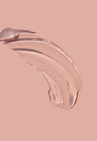 Kirkas, kiiltävä geelimäinen tahra, jossa on sileä pinta ja hienovaraisia kuplia, tasaisella haalean vaaleanpunaisella taustalla.