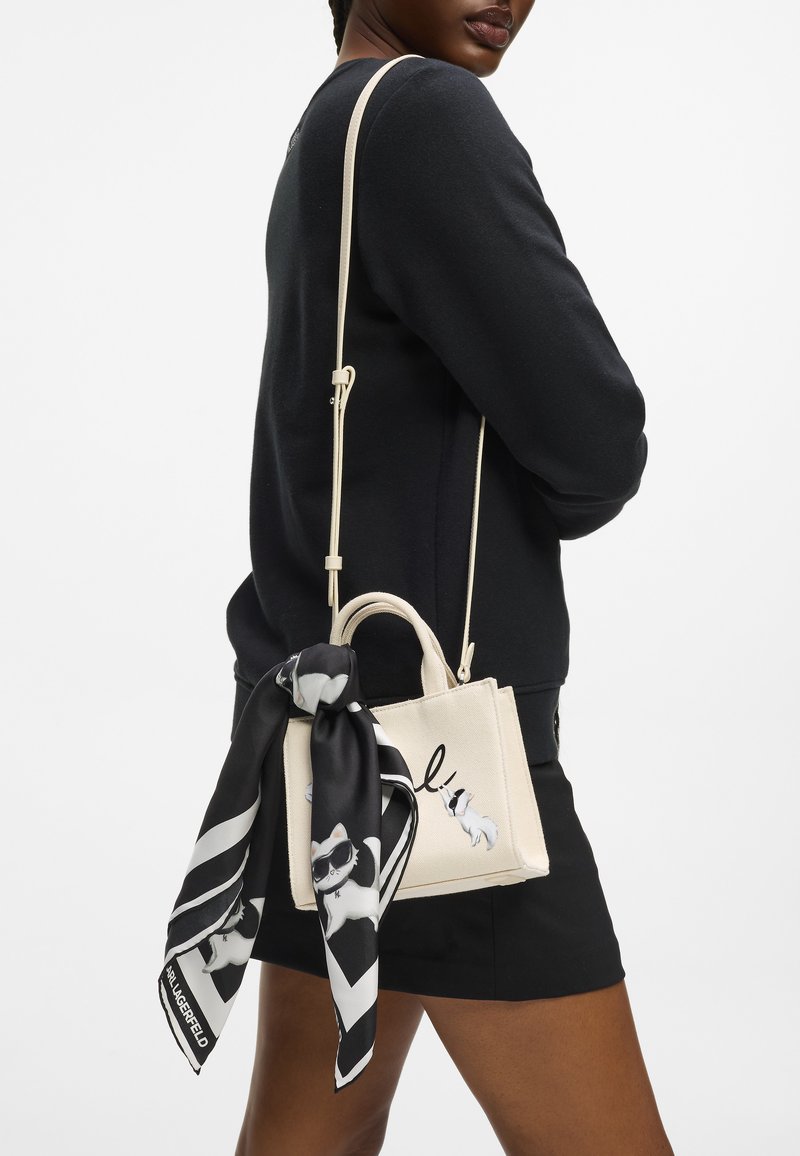 Personne portant une tenue noire avec un sac fourre-tout crème orné d'un imprimé de chat en noir et blanc et une écharpe assortie nouée à la poignée du sac.