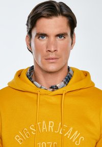 Żółta bluza z kapturem z haftem "BIG STAR JEANS 1970" z przodu, noszona na niebieskiej koszuli w kratę z guzikami. Gładka tekstura, casualowy design.