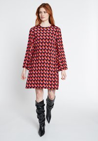 Rood-zwart geometrisch patroon jurk met een losse pasvorm, lange mouwen en een ronde halslijn, gecombineerd met kniehoge zwarte laarzen.