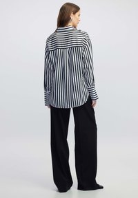 Schwarz-weiß gestreiftes Langarmhemd mit Kragen, versehen mit einem abgerundeten Saum und weitem schwarzen Hosen. Taillierte Passform, glatte Stofftextur.