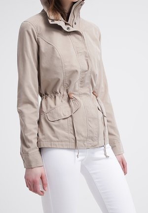 Vrouw draagt een beige jack met ritssluiting, hoge kraag en taillekoord, gecombineerd met een witte broek voor een effen achtergrond.