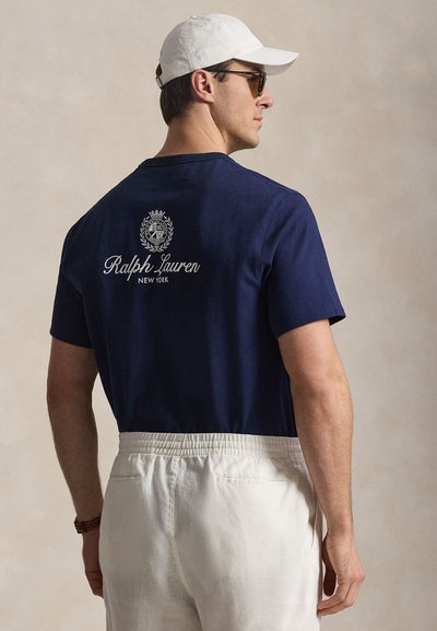 Άνδρας με μπλε ναυτικό t-shirt Ralph Lauren, λευκό παντελόνι, λευκό καπέλο και γυαλιά ηλίου, γυρισμένος ελαφρώς στο πλάι μπροστά σε απλό φόντο.