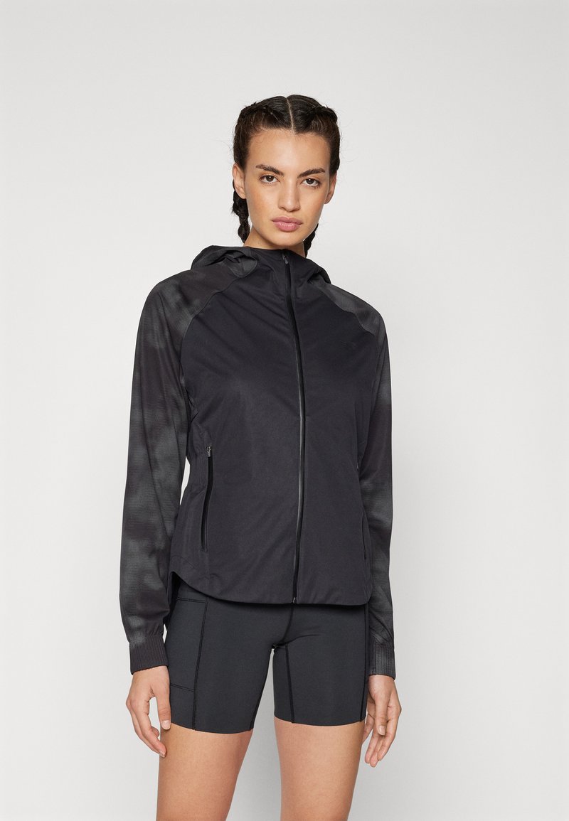 Schwarze Kapuzenjacke mit vollem Reißverschluss, hat gemusterte Ärmel und besteht aus einem leichten Material. Getragen mit schwarzen Shorts.