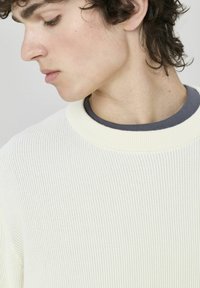 Pull en maille blanche à côtes avec une finition texturée et un col rond, superposé sur une chemise intérieure marine, avec des détails de couture subtils.