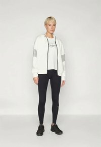 Biała bluza z zamkiem błyskawicznym w czarne paski, zestawiona z białą koszulką i czarnymi legginsami. Czarne buty sportowe dopełniają całość.