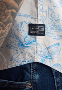 Witte shirt met een blauw bloemenpatroon en een label met de tekst "Petrol Ind." Blauwe denim jeans zichtbaar aan de onderkant met een metalen knop.