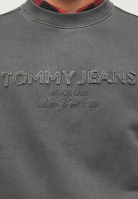 Sudadera gris con el texto bordado "TOMMY JEANS", "SINCE 1985" y "New York City" en una textura suave y con cuello acanalado.