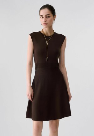 Femme portant une robe sans manches marron foncé avec des côtes à la taille, accessoirisée de colliers en or superposés et de boucles d'oreilles, debout devant un fond uni.