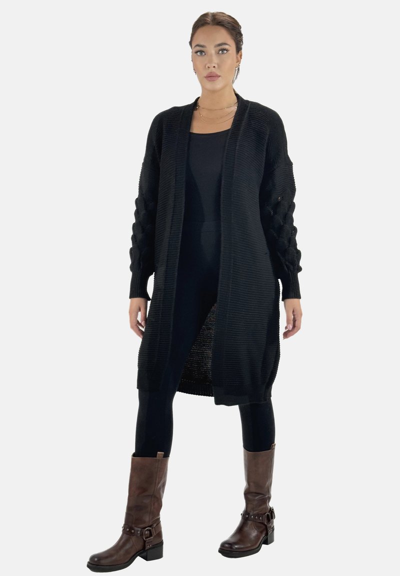Svart stickad cardigan med texturerade, kabel-stickade ärmar och öppen front, kombinerad med svarta leggings och bruna stövlar som når till mitten av vaden.