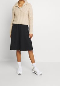 Beige pletený svetr s polozipem, kombinovaný s černou prošívanou sukní ve tvaru A. Nosený s bílými teniskami a kotníkovými ponožkami.