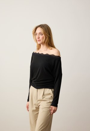 Kobieta z długimi blond włosami, ubrana w czarną bluzkę odsłaniającą ramiona i beżowe spodnie, stojąca na tle jednolitego jasnego tła.