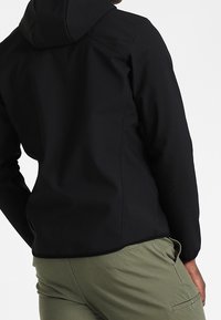 Giacca nera con cappuccio e finitura liscia e opaca. Presenta polsini elasticizzati e un design aderente. Abbinata a pantaloni cargo verde oliva.