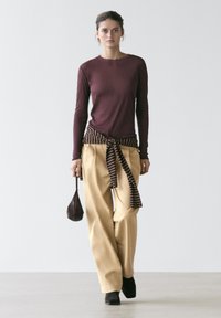 Femme marchant en pantalon ample beige, haut à manches longues marron, pull rayé noué à la taille, chaussures noires et tenant un petit sac marron.