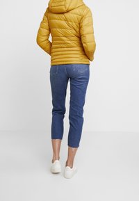 Chaqueta de plumas amarilla con capucha, paneles texturizados; vaqueros de mezclilla recortados azules con dobladillo deshilachado; zapatillas blancas. Vista desde la parte de atrás.