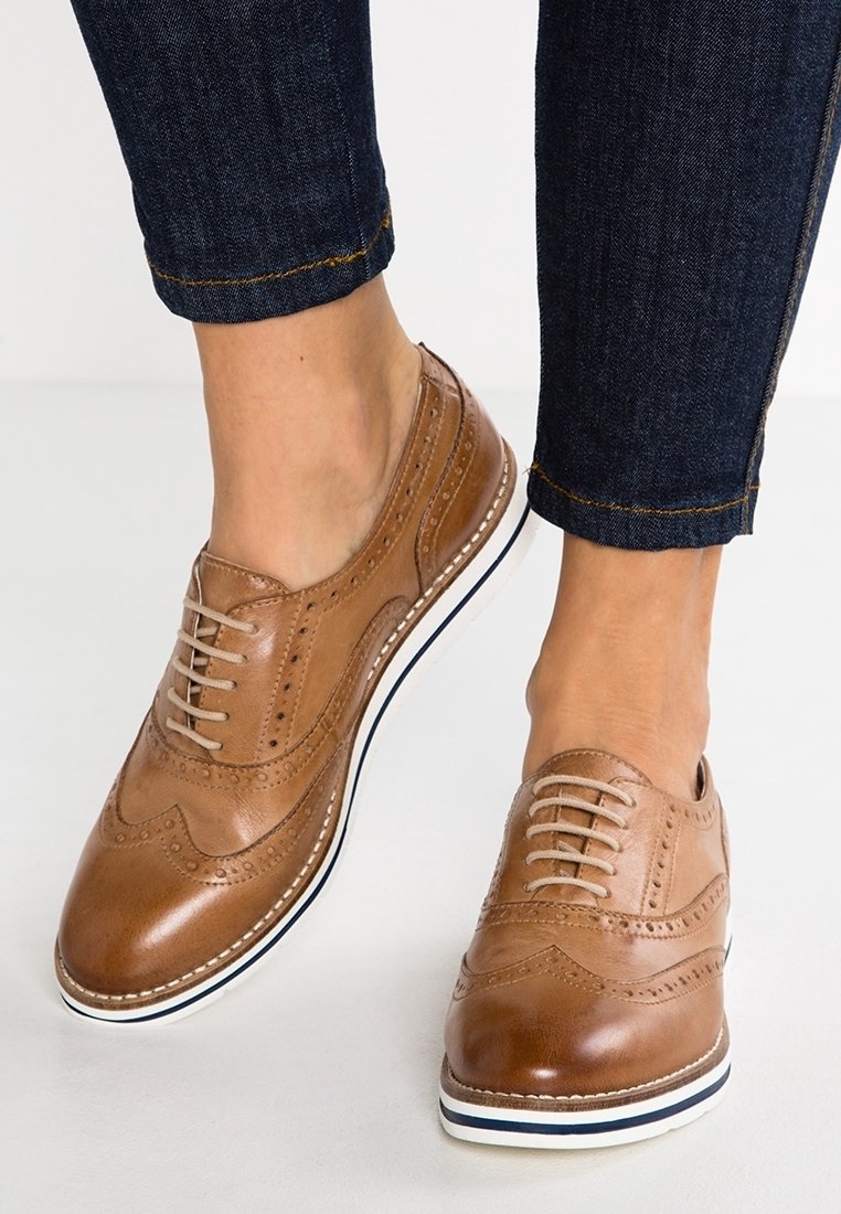 Chaussures brogues en cuir brun clair avec des lacets beige, portées avec un jean en denim bleu foncé, montrant des coutures détaillées et des semelles blanches avec une bande marine.